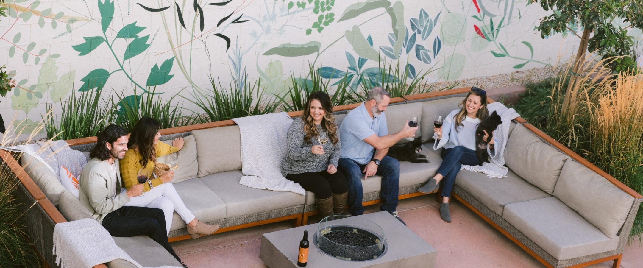 5 people sitting on u-shaped patio furniture around a fire pit drinking wine. One woman is holding a small black dog.