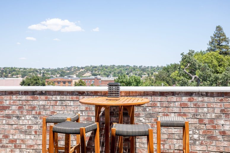 A hightop table and 4 stools along a brick wall on a roof top. Greenery view in the distance.