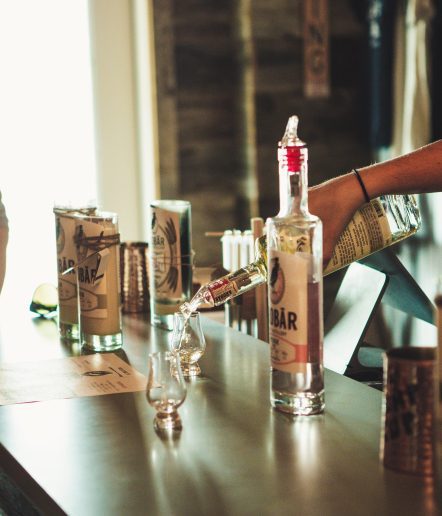 a counter with jar candles, a bottle of liquor , 2 cordial glasses and someone pouring liquor into one of the glasses