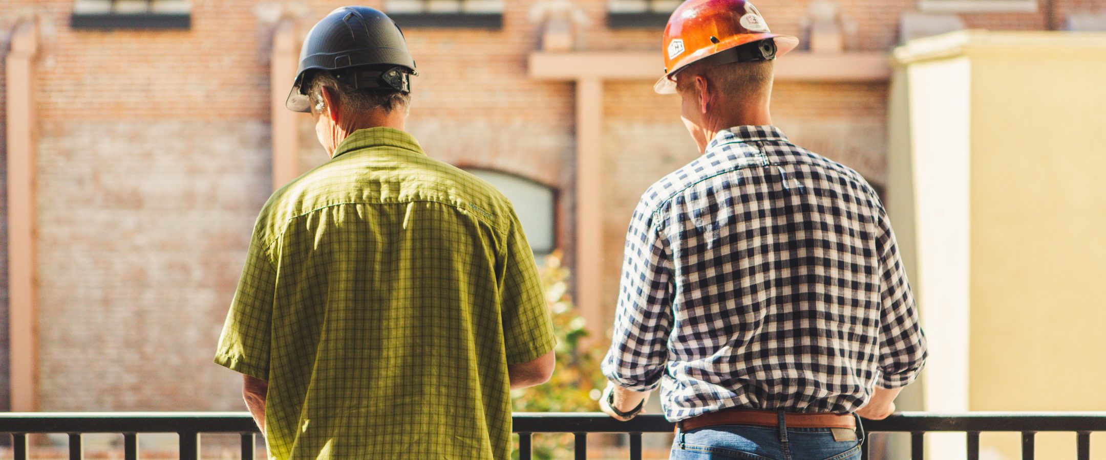 two men in plaid shirts wearing hard hats standing at a railing looking at a brick building