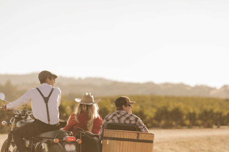 A couple in a sidecar being driven by a vineyard. The woman has blond hair and is wearing a cowboy hat. The man is wearing a baseball cap. The driver is wearing a whit shirt and suspenders.