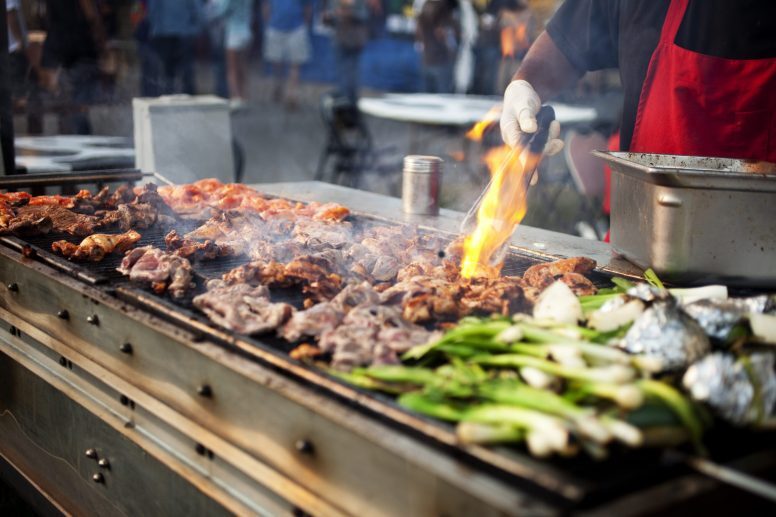 Chicken being flame broiled on a large grill with a man wearing a red apron and gloves holding tongs.