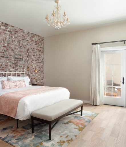 A bed with a bench at the foot of it and chandelier lighting above it. A brick wall with bedside lighting is visible behind the headboard.