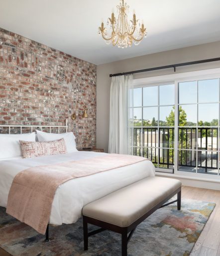 A king bed with white blanket and pink accents with a bench at the foot of the bed, Large glass doors overlook greenery