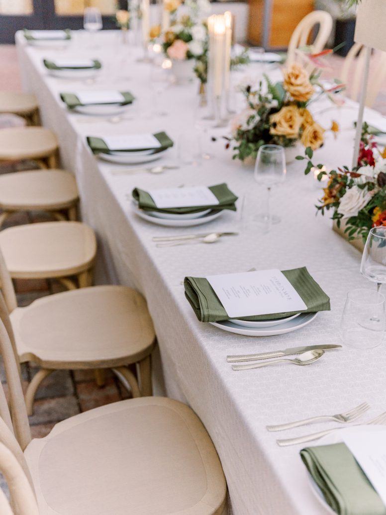 Table set with white linens, green cloth napkins, menus on top of plate, and color floral arrangements.