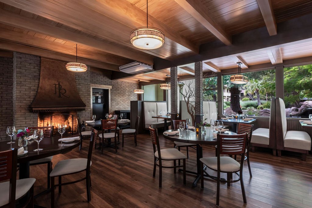A photo of the dining room at Paso Robles inn. Wood floors, tables, a fireplace and a wall of windows fill the space.