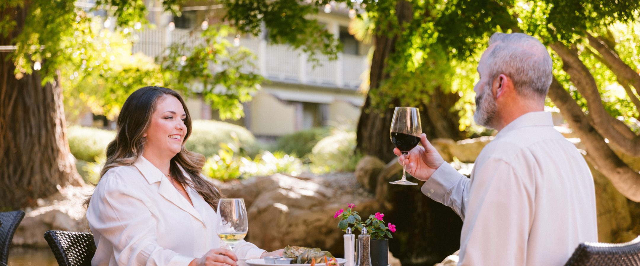A couple seated at a table outside. The man hold up a glass of wine. The woman is in a white shirt.