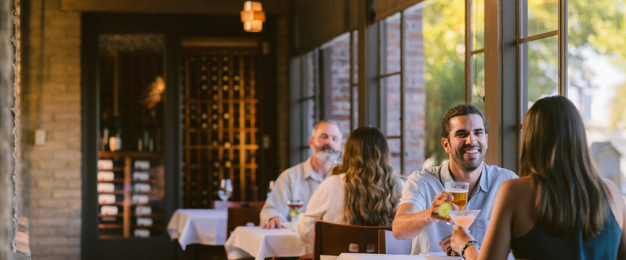 People on a restaurant seated at tables with white tablecloths along a row of windows.