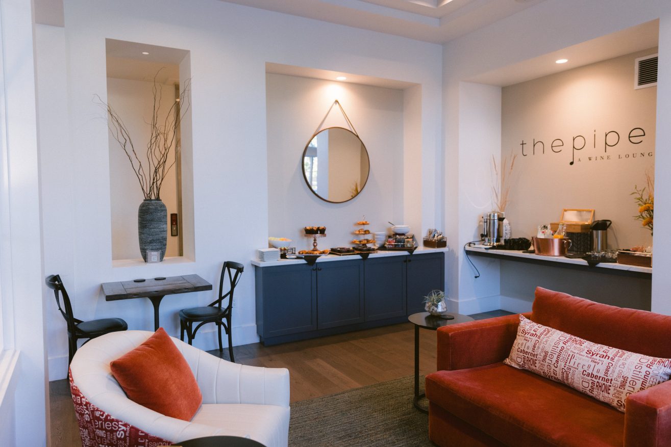 A room with sofa, chair, table and 2 chairs. There is a coffee station and a breakfast buffet with muffins set up along the wall behind the seating area.