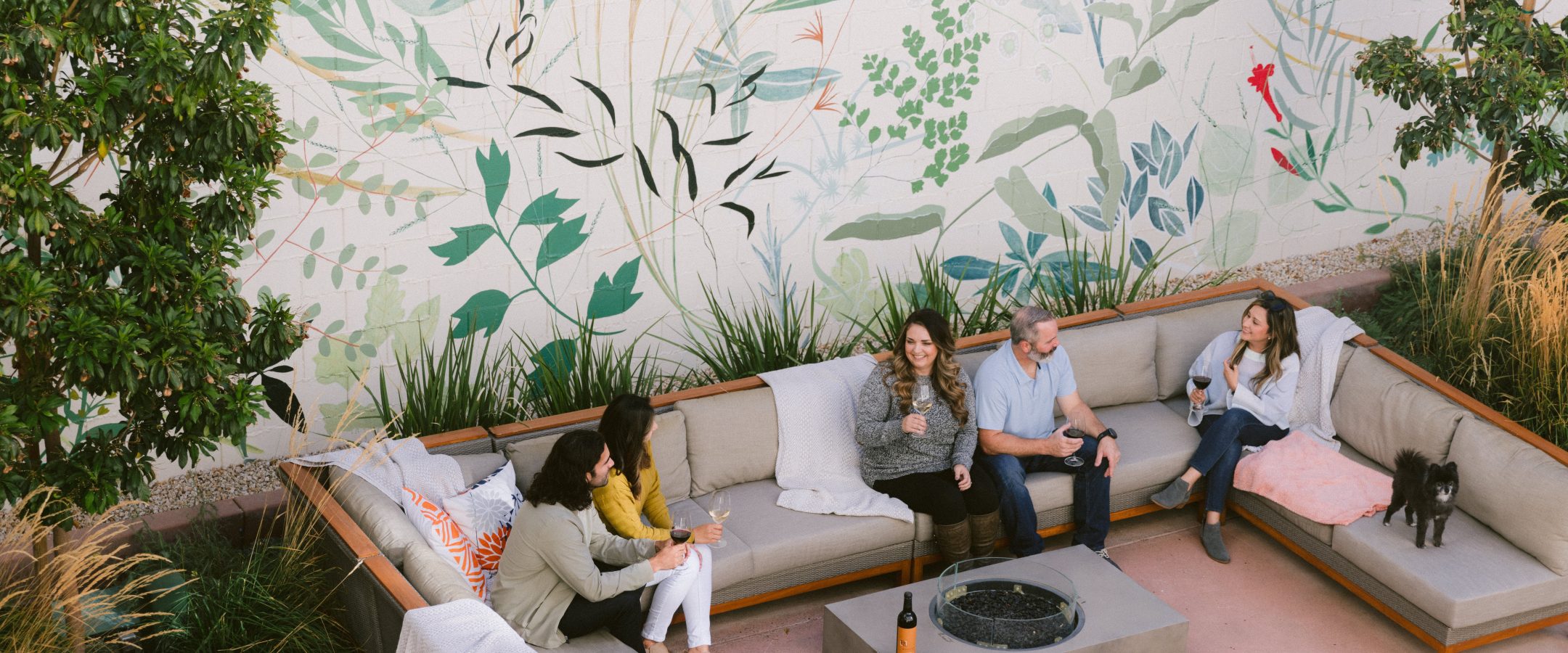 5 people sitting on u-shaped patio furniture around a fire pit drinking wine. One woman is holding a small black dog.