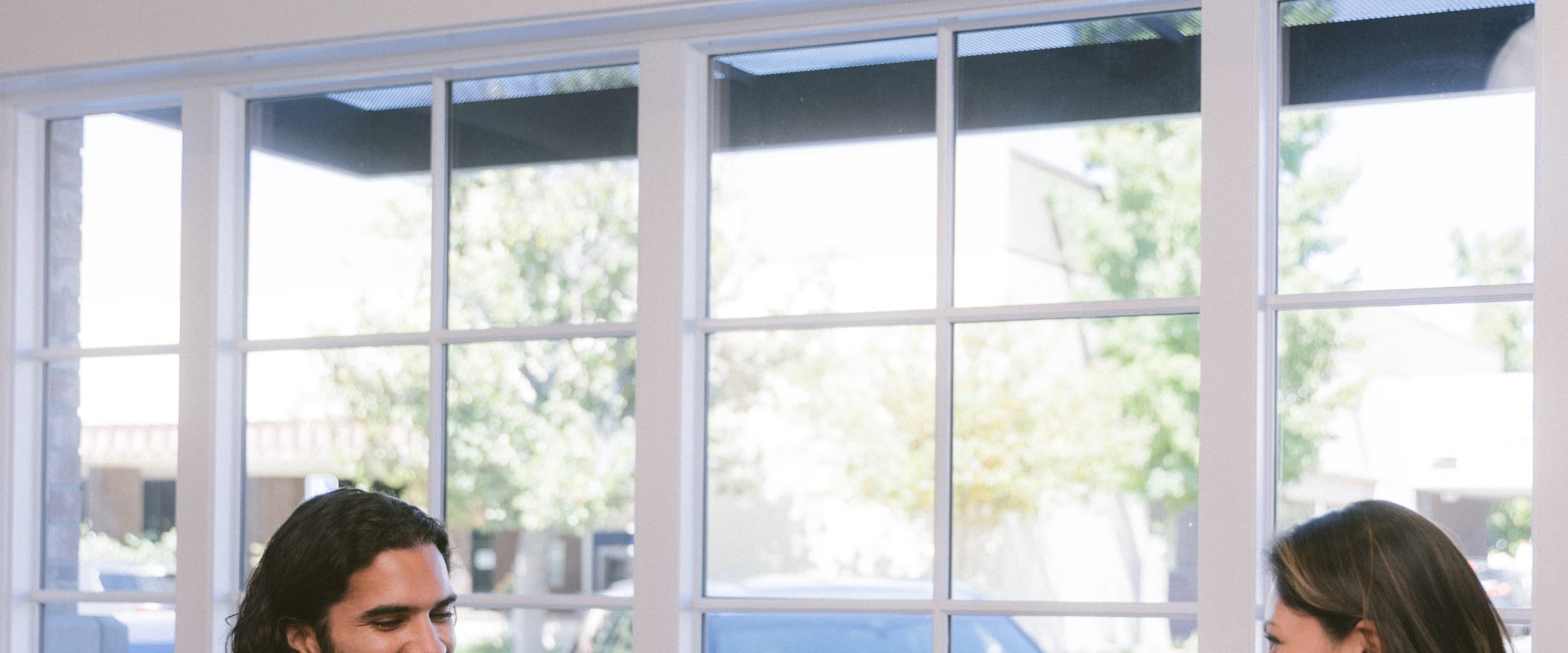 A man with a beard sitting at a table by a window. There is a cheese and fruit platter on the table too.