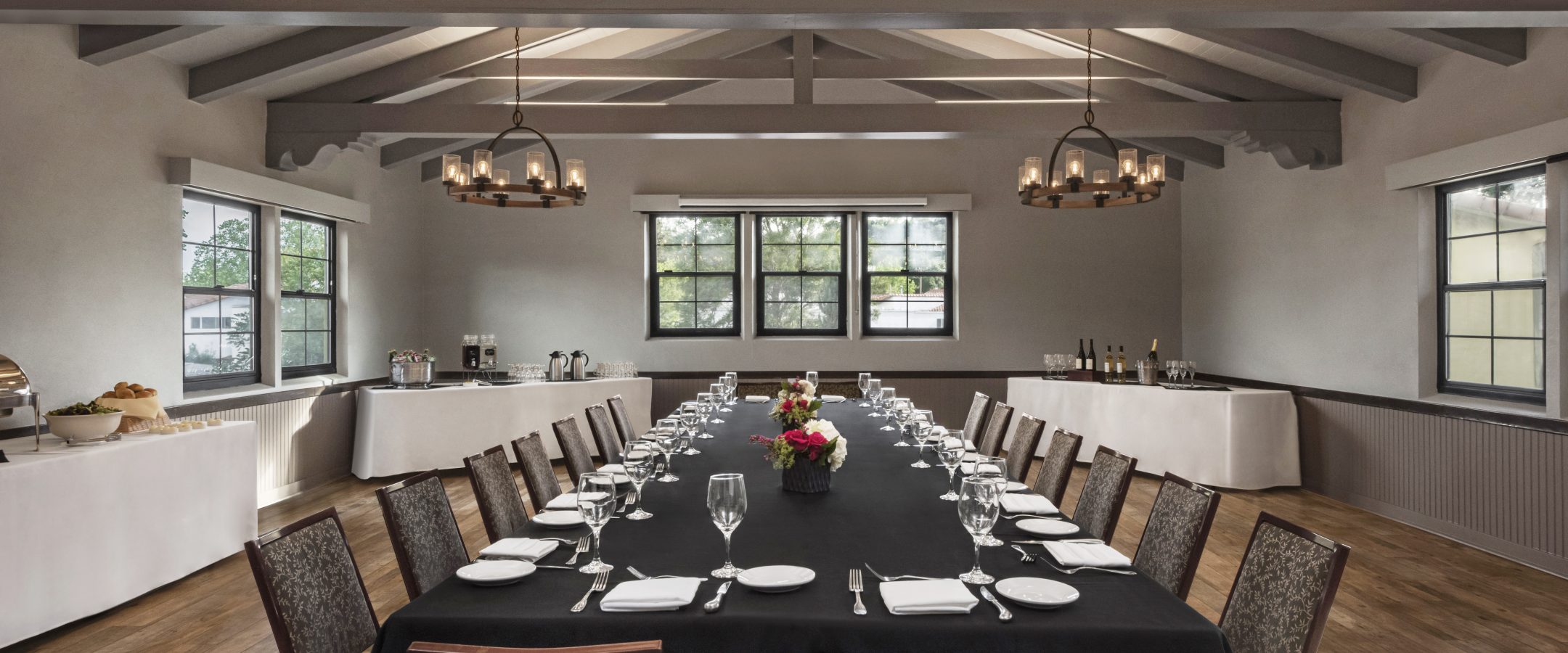 A dining room with a large table in the center of the room, surrounded by dining chairs with white plates, cutlery, and empty wine glasses.