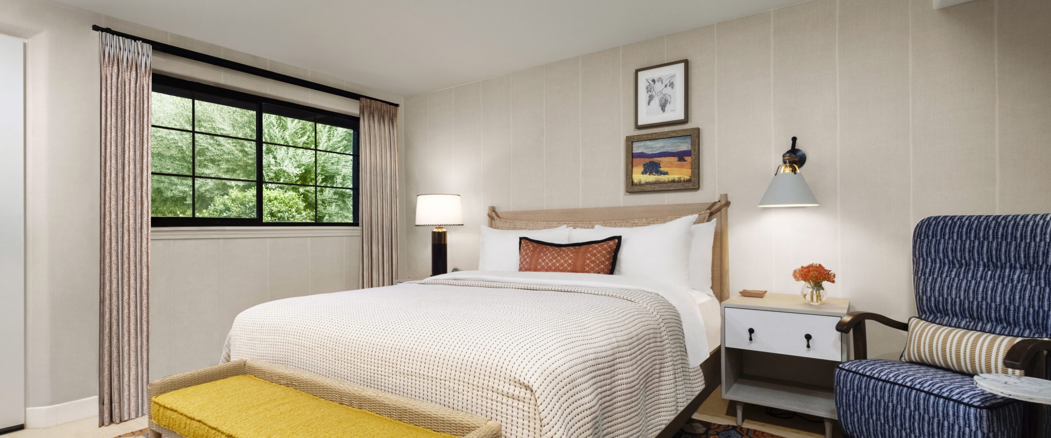 A bed with a bench at the foot of it, with side tables on each side with lighting and a blue armchair beside the bed.  
