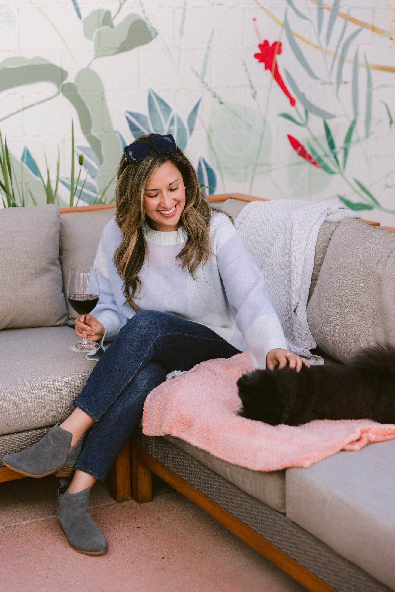 A woman sits outside on a sofa with her black dog next to her on the sofa. The dog is on a blanket.