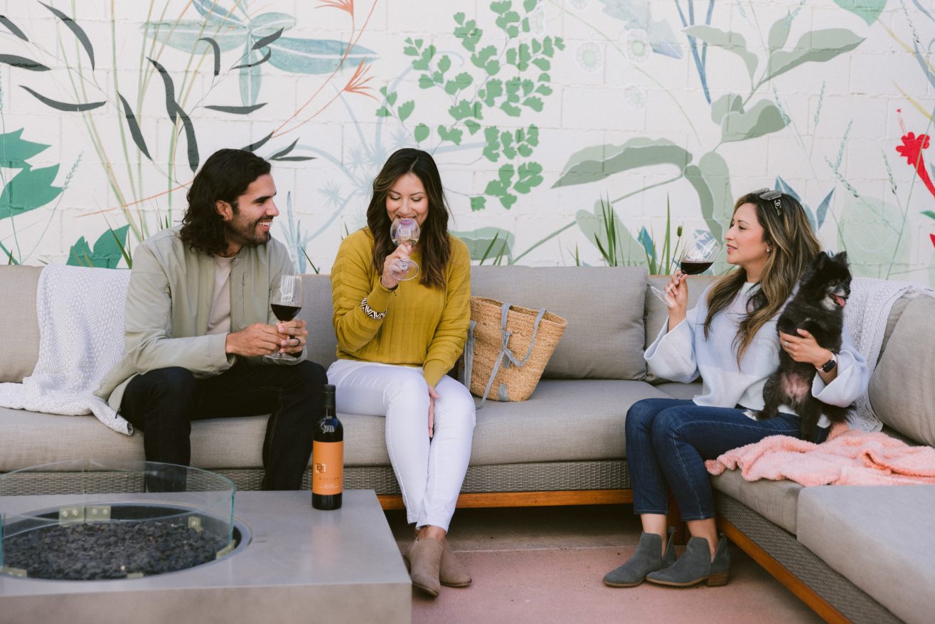 A man, two ladies and a small dog sitting out while enjoying glasses of wine together.