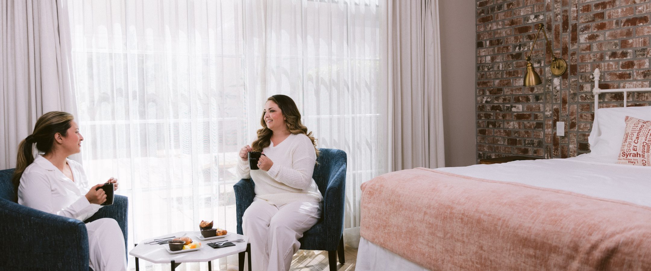 Two happy women seated next to a bed and by the window, enjoying tea and pastries.