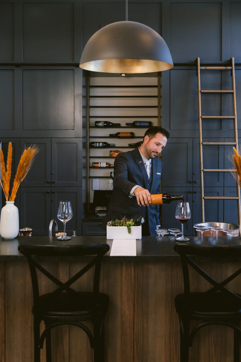 a sommelier pouring wine with a wine refrigerator behind him