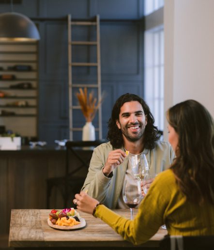 A man in a green jacket and lady in yellow wine tasting and eating a charcuterie board.