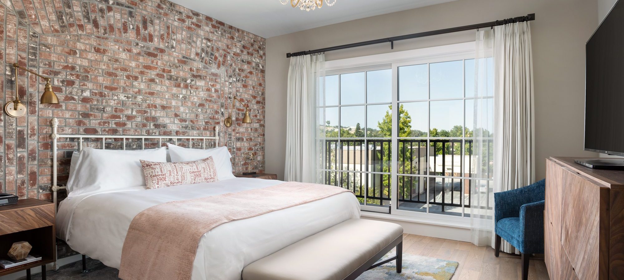 A king bed with white blanket and pink accents with a bench at the foot of the bed, Large glass doors overlook greenery