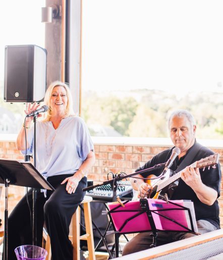 Two musicians - a lady singing into a microphone and a man playing a guitar on a rooftop deck.