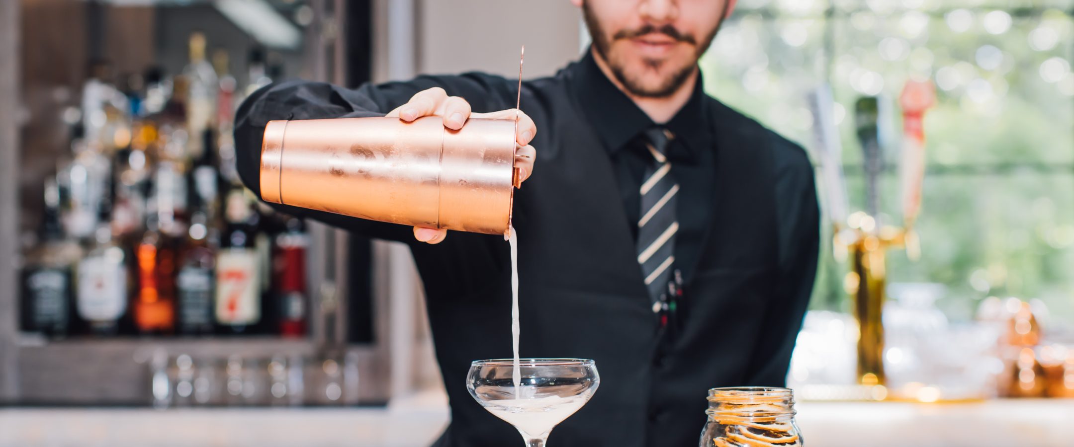 The torso of a bartender shown pouring a drink from a shaker into a martin glass.