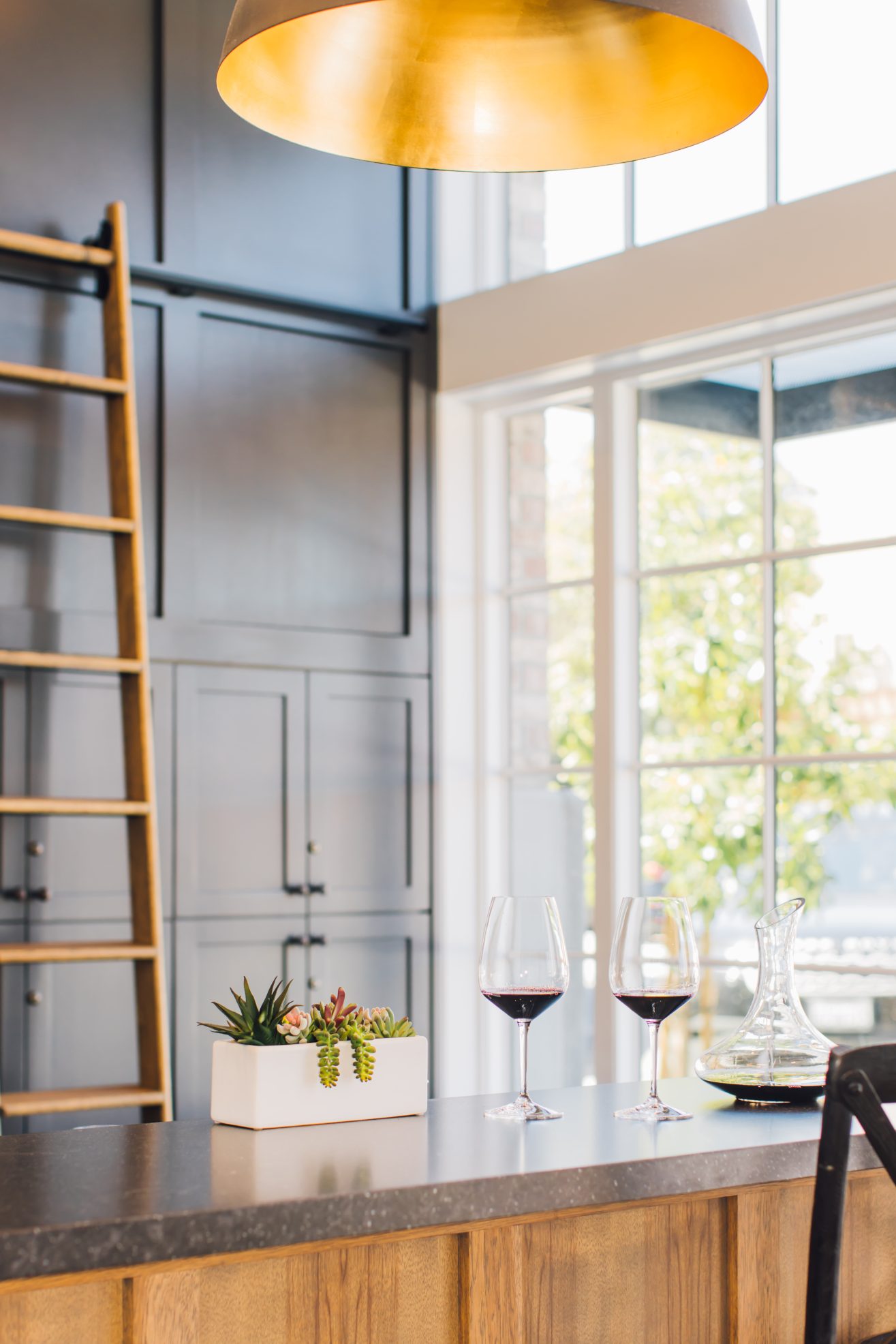 A wine decanter and two glasses with red wine on top of a bar. The bar is next to large windows. There is a library ladder along the back wall.