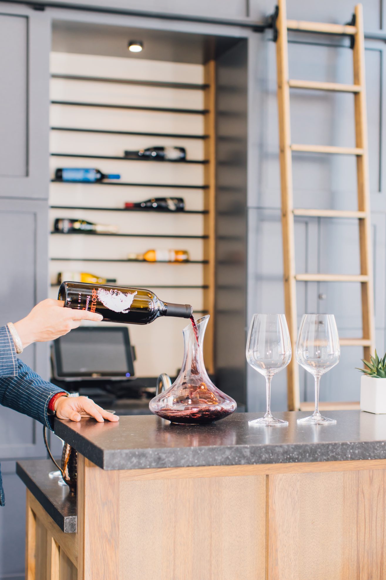 A close up of wine being poured from a decanter into wine glasses. The is a rack with wine bottled on the wall behind the bar.