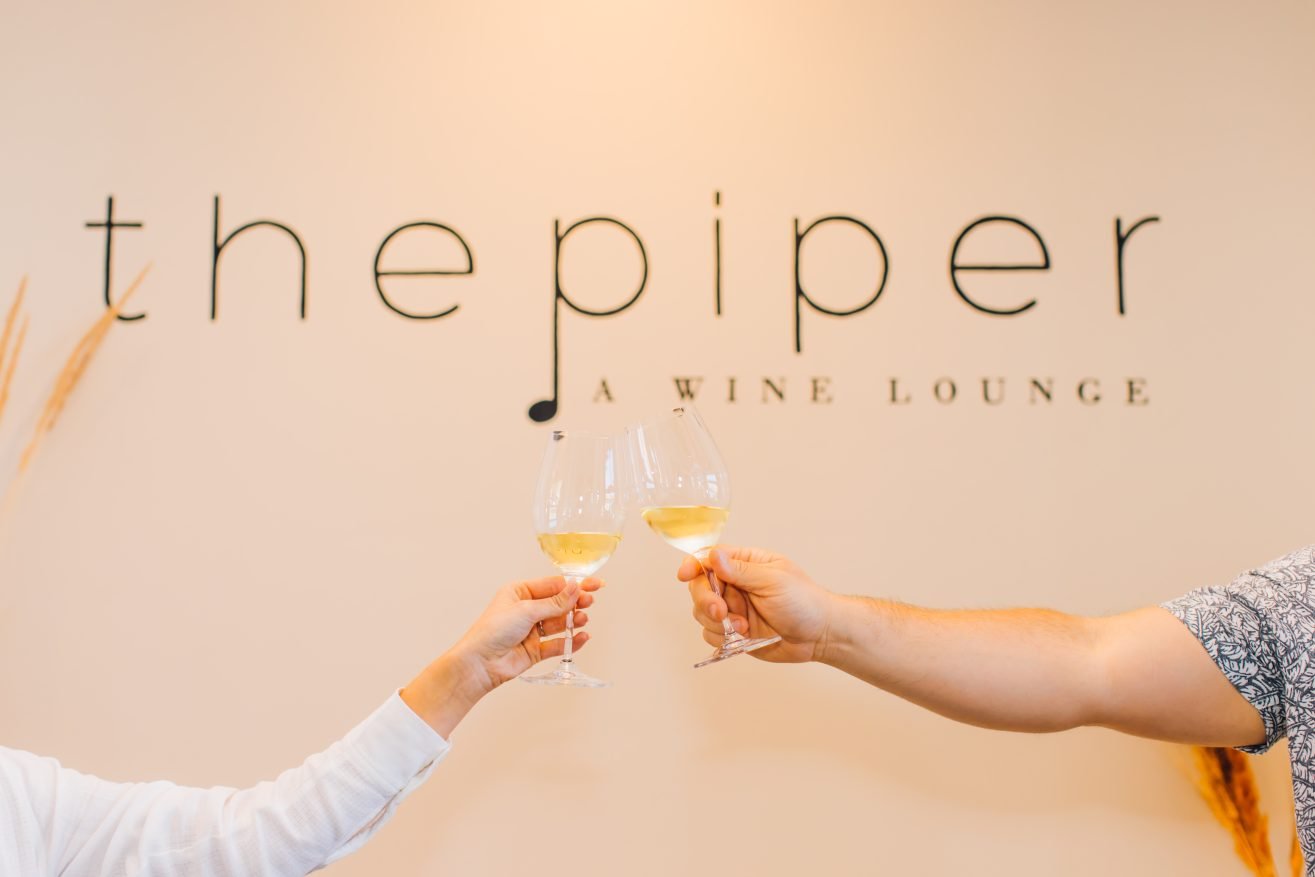 The Pieper Lounge sign on the wall with two people's arms in the foreground raising a glass of wine.