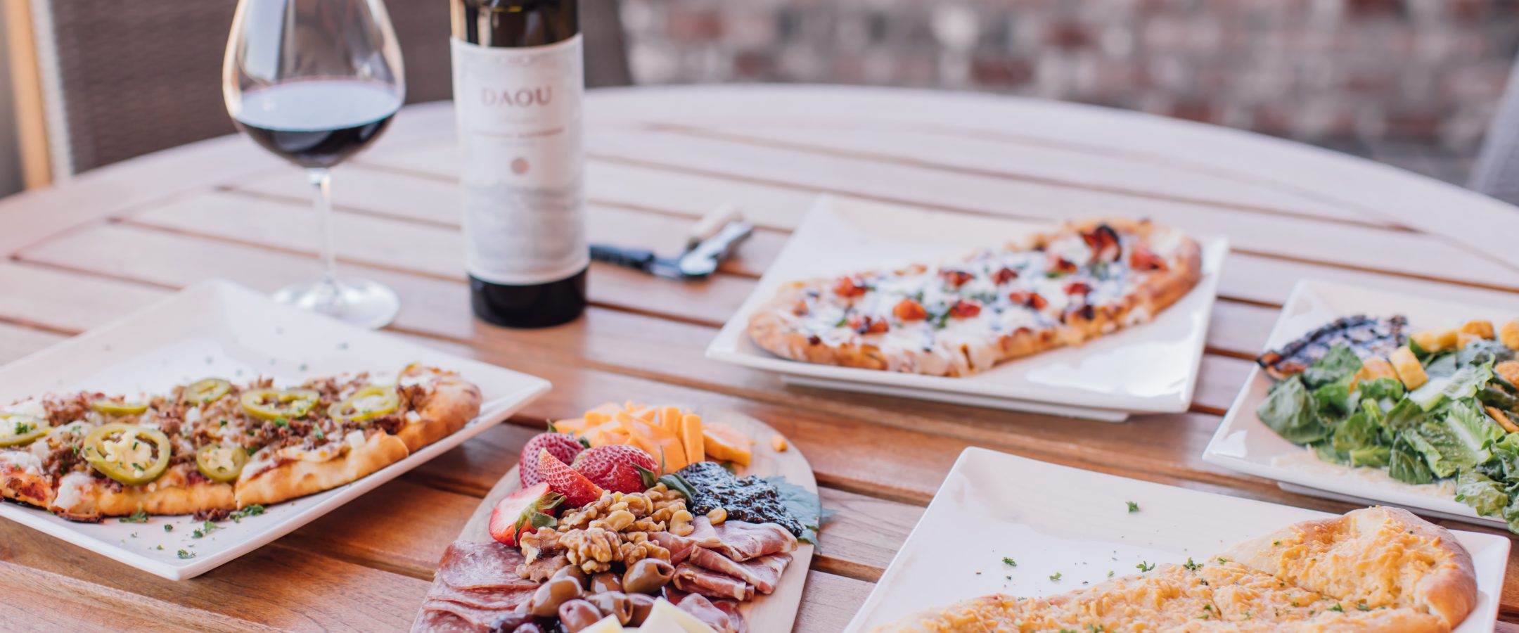 A patio table with a variety of flatbread pizzas, and a bottle of red wine.