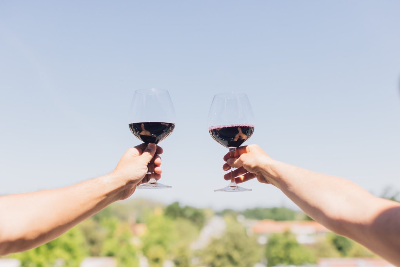 Two glasses of red wine being raise with greenery in the distance.