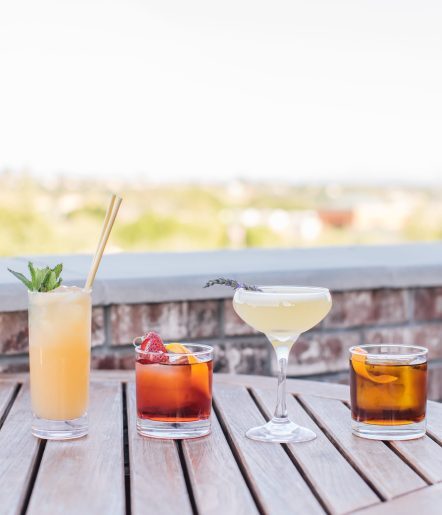 A variety of cocktails in different glasses on a wooden table on a rooftop. The view of greenery is behind the glasses.