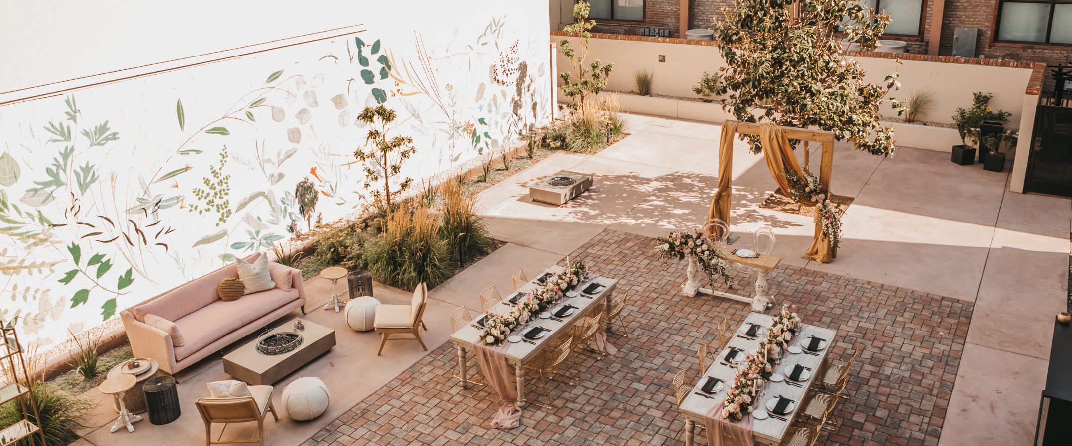 An aerial view of the courtyard with a flora mural and brick wall. The courtyard has t wo dining tables set for dinner and a seating area with a fire pit.  