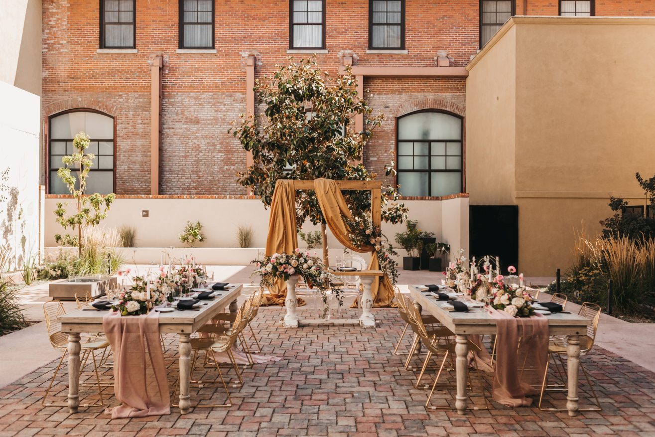 A courtyard with two wooden dining tables and gold chairs. The tables are set with bouquets and plate ware. There is an alter in front of a tall green tree with a brick building behind.