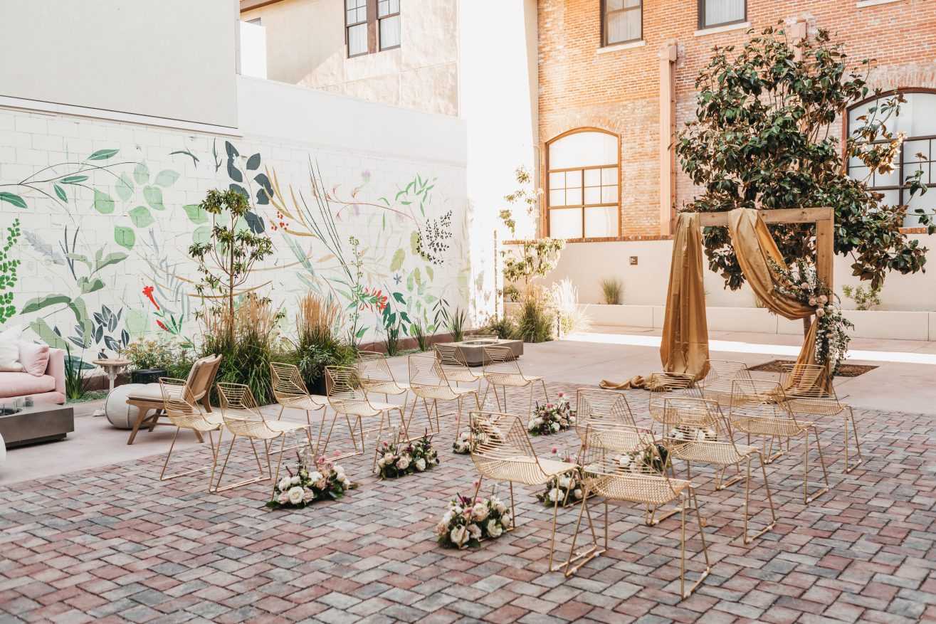 A courtyard with a floral mural on one wall set up for a wedding with 22 aisles of gold chairs, and alter in front of a tall green tree