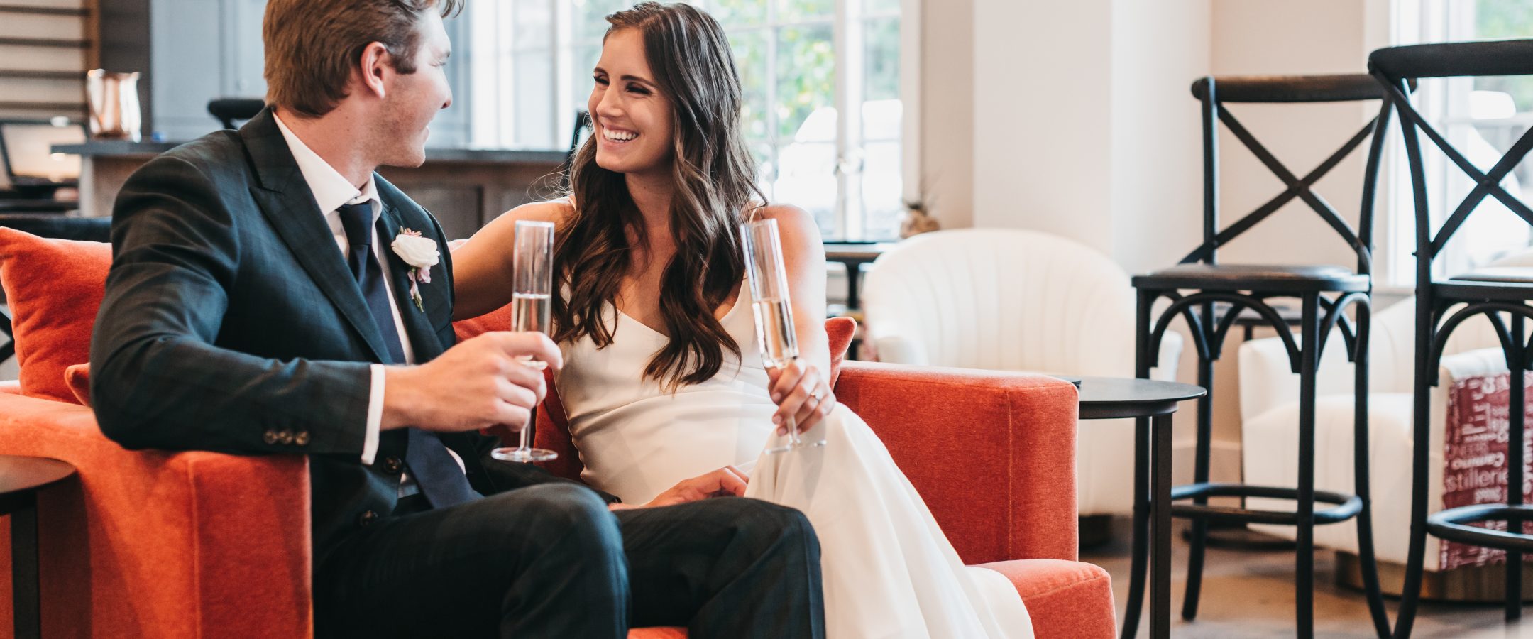 A bride and groom sit on a sofa smiling at each other. There are large windows behind them.