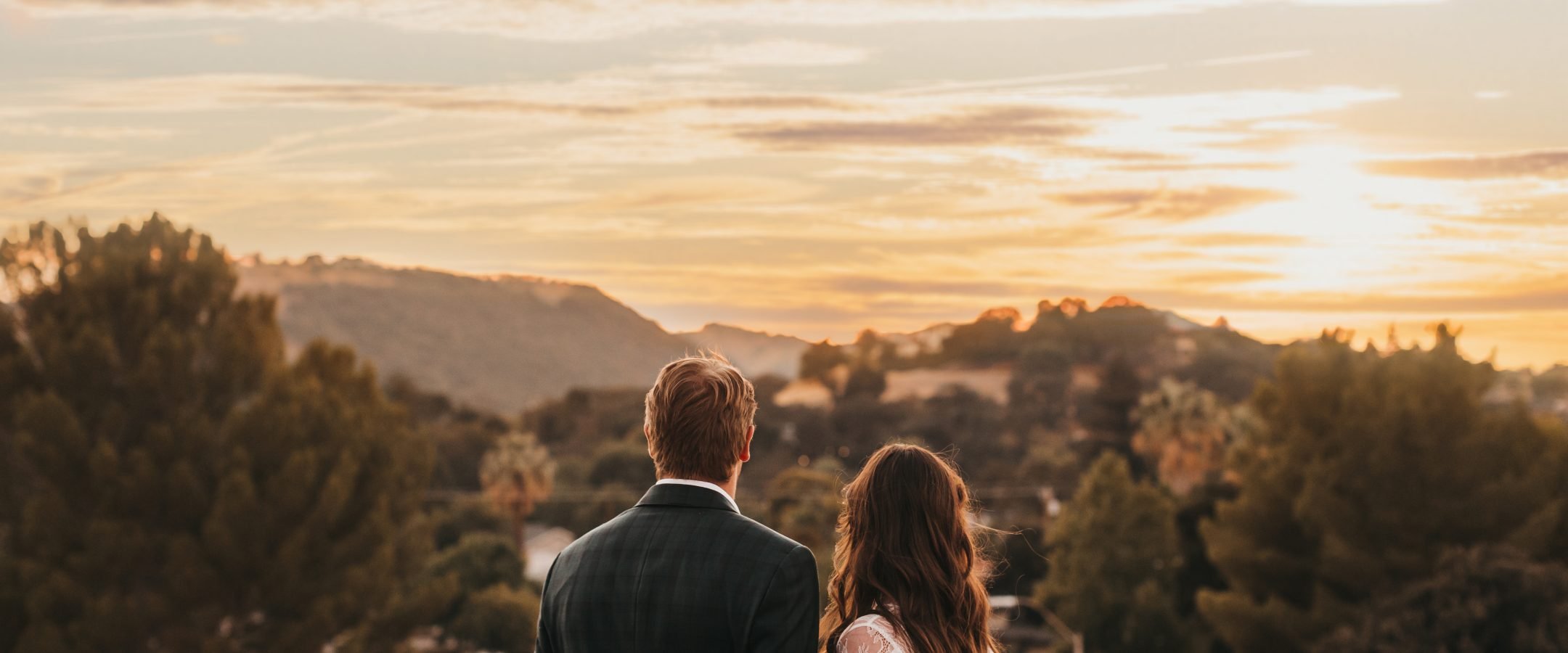 A bride and groom look out to the setting sun on the horizon from a roof top bar.