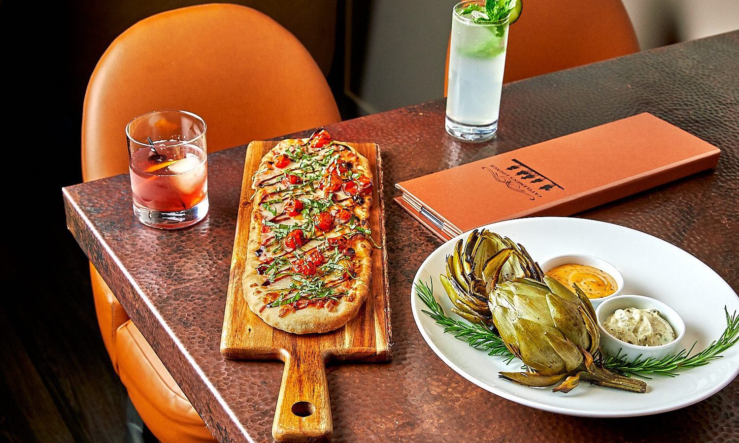 A close up of a table with a cocktail, a flatbread pizza, and grilled artichoke on top of it.