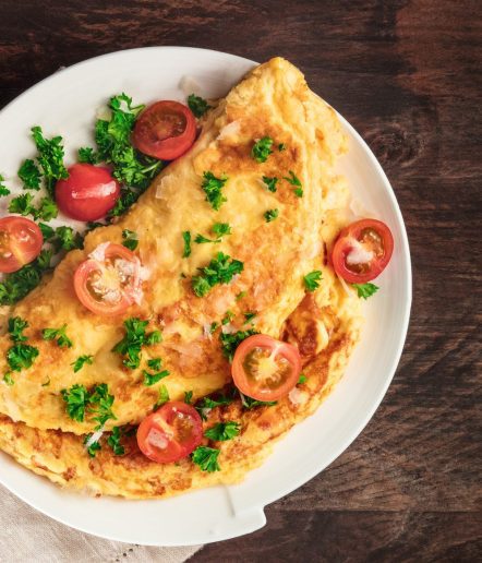 A photo of an omelet with cherry tomatoes, parsley. and grated cheese, shot from above on a rustic wooden texture with a place for text