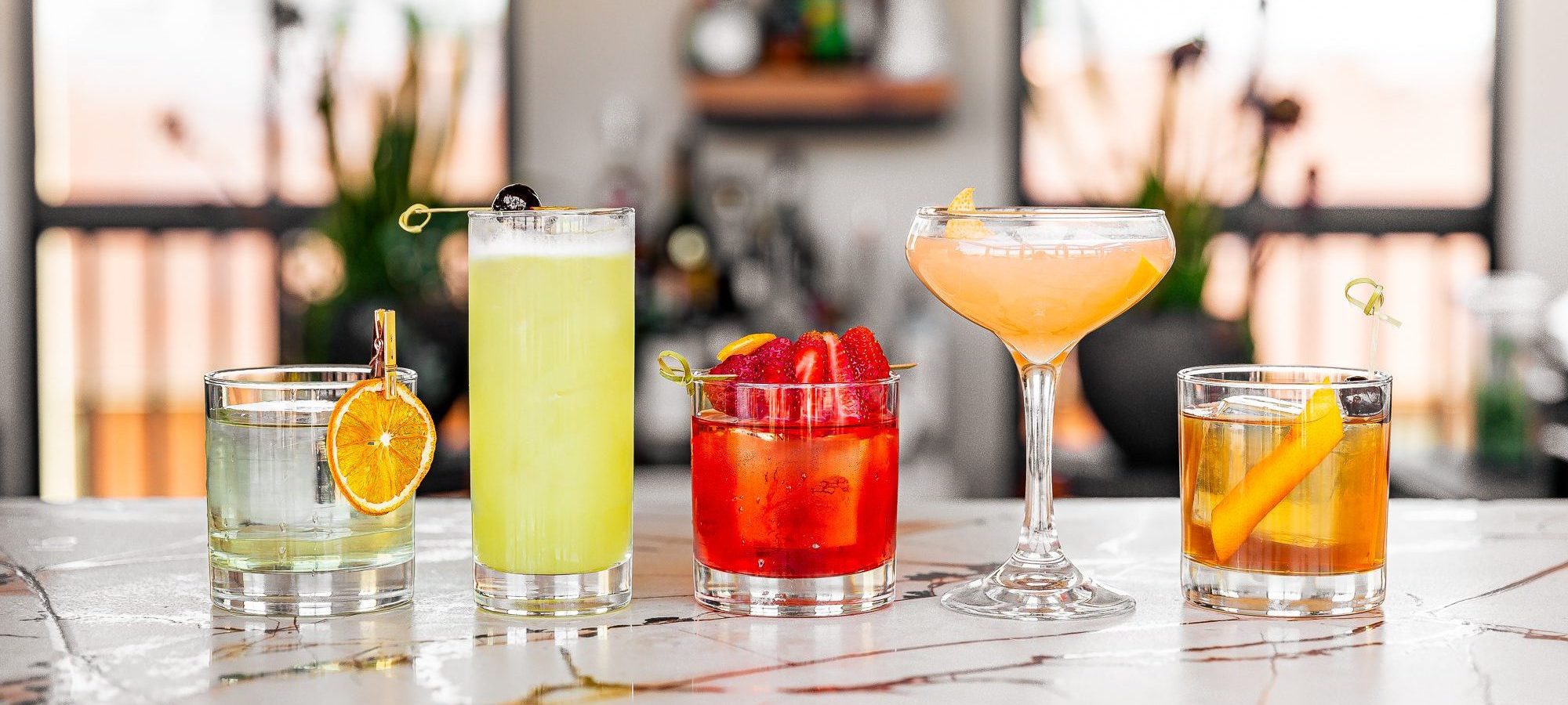 A variety of cocktails on a bar. Each is a different color in a different glass.