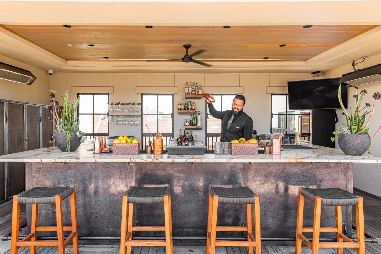 A bartender stands behind a bar shaking a cocktail. THree barstools are in the foreground