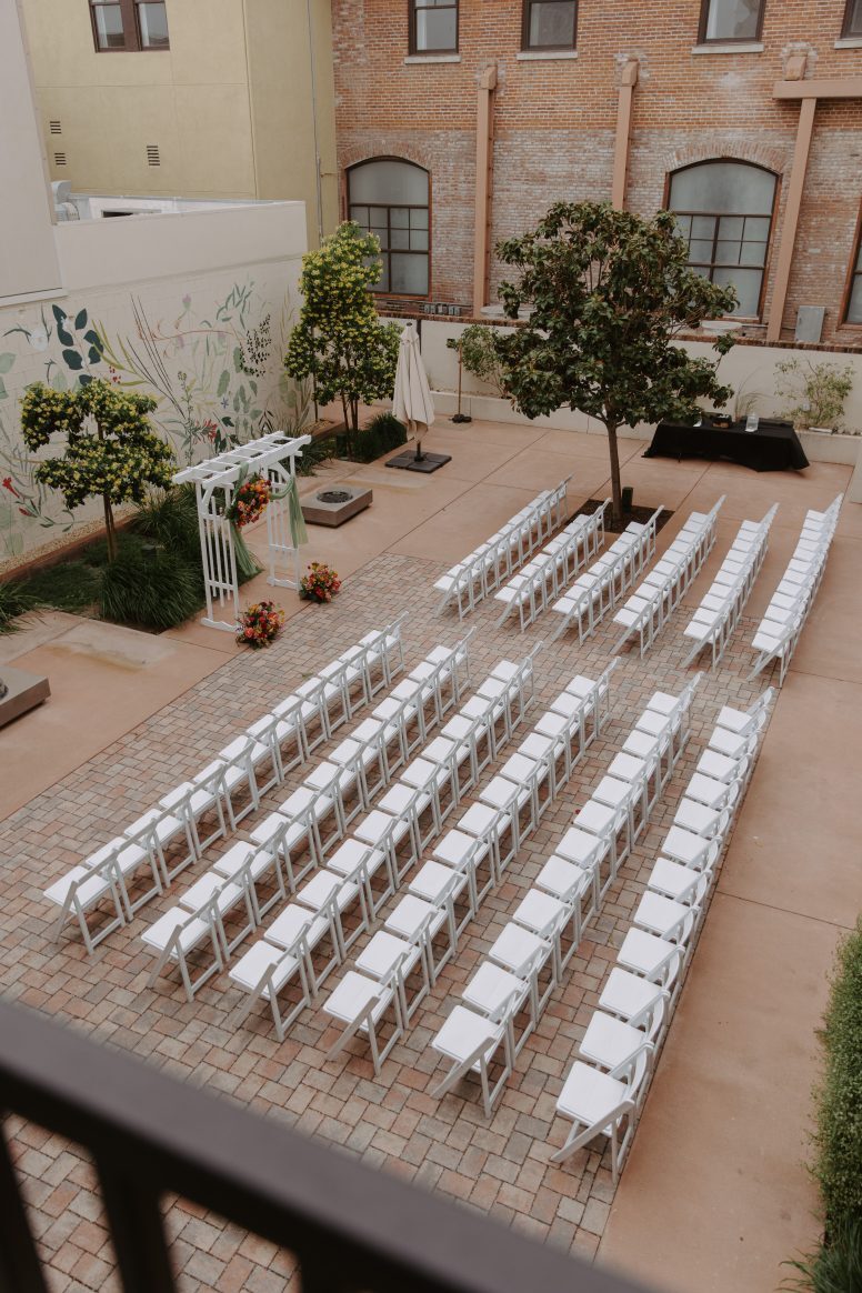 A setup of white folding wedding chairs in front of wedding arch and colorful mural in a courtyard.