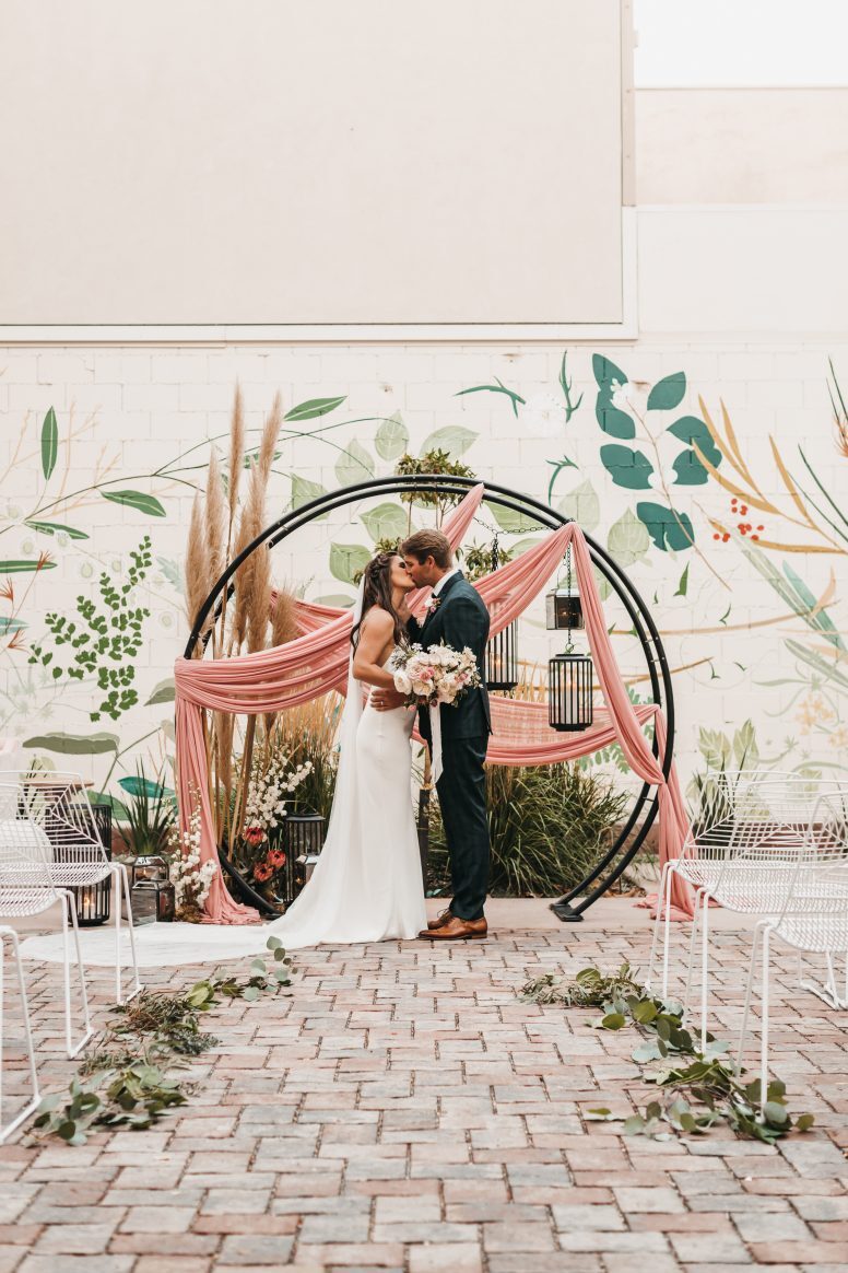 A bride in white and groom in black kissing in front of circular altar arch and colorful mural.