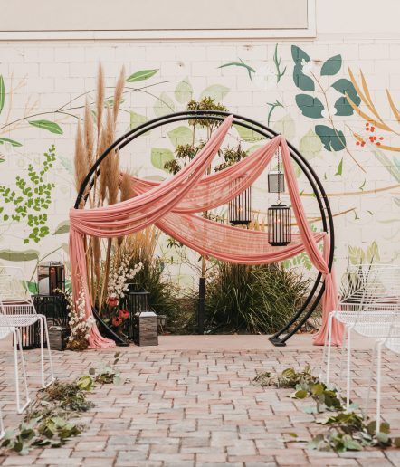 An outdoor circular altar adorned with floral arrangements, surrounded by white chairs facing it, with a large mural wall in the background.