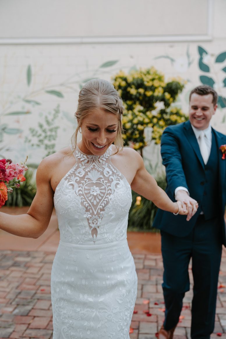 A bride in white holding bouqet in one hand and groom in blue holding other hand smiling in front of a colorful mural.