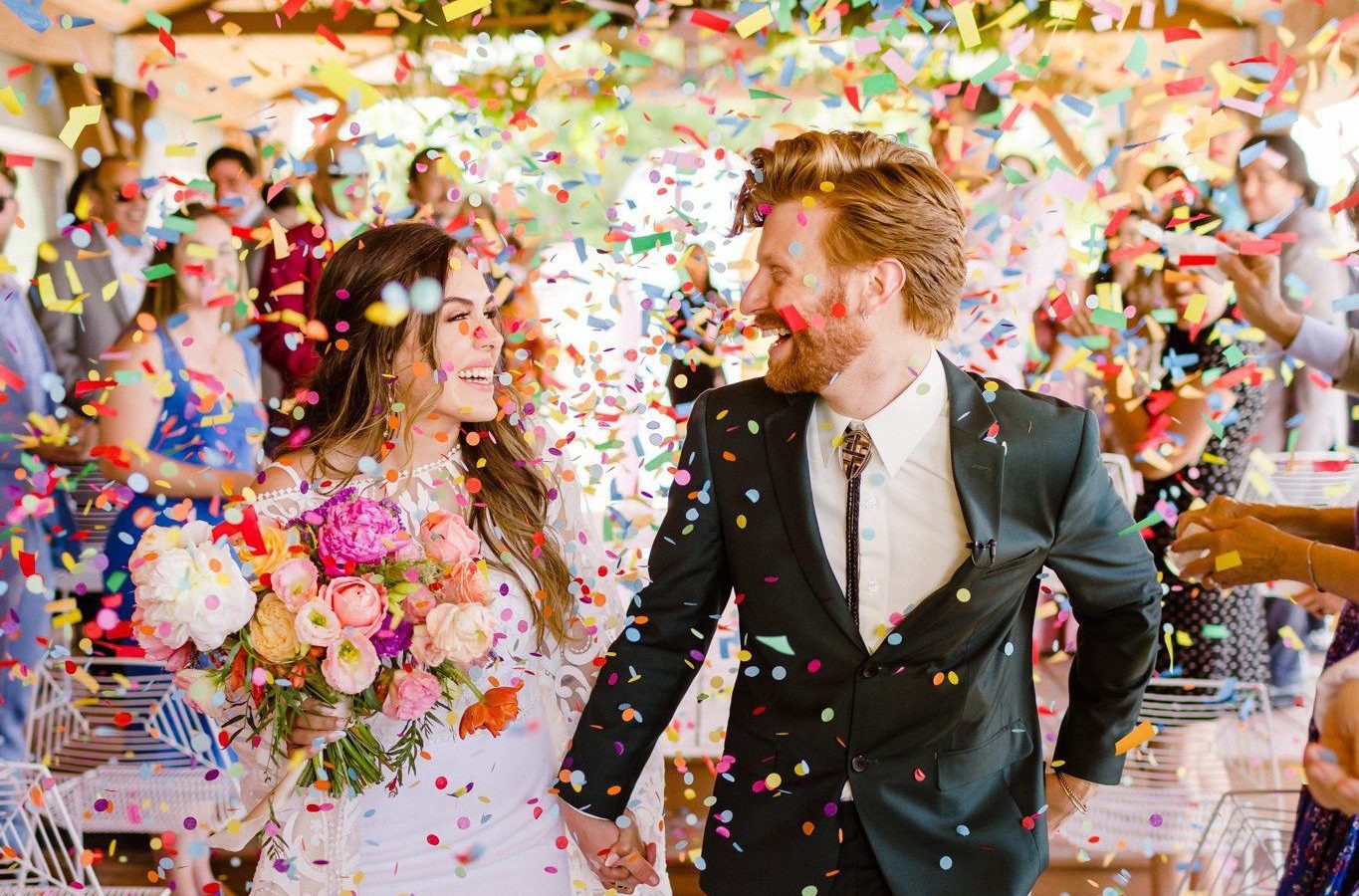 A bride in white and groom in black with a bolo tie with colorful confetti in the air.