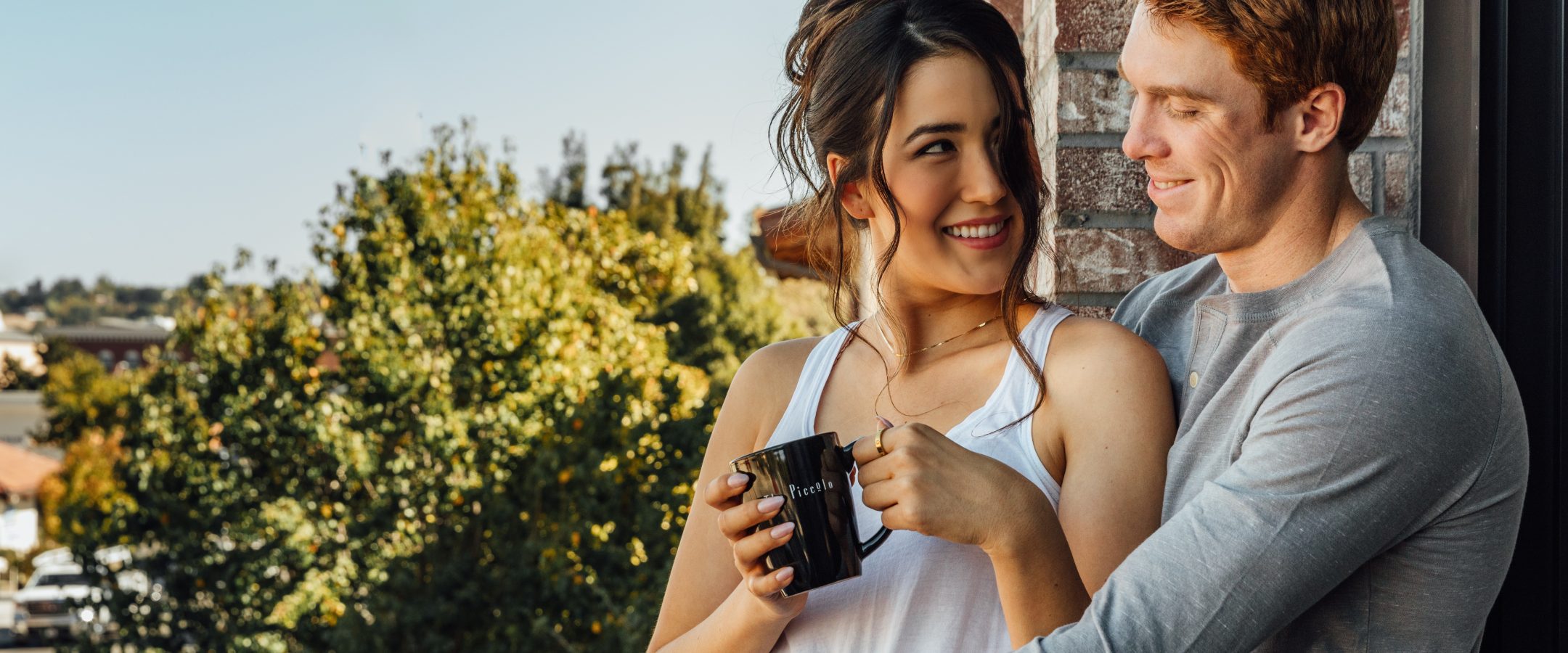 A cheerful man and woman standing outside and looking at each other with piccolo branded coffee mugs in their hands.