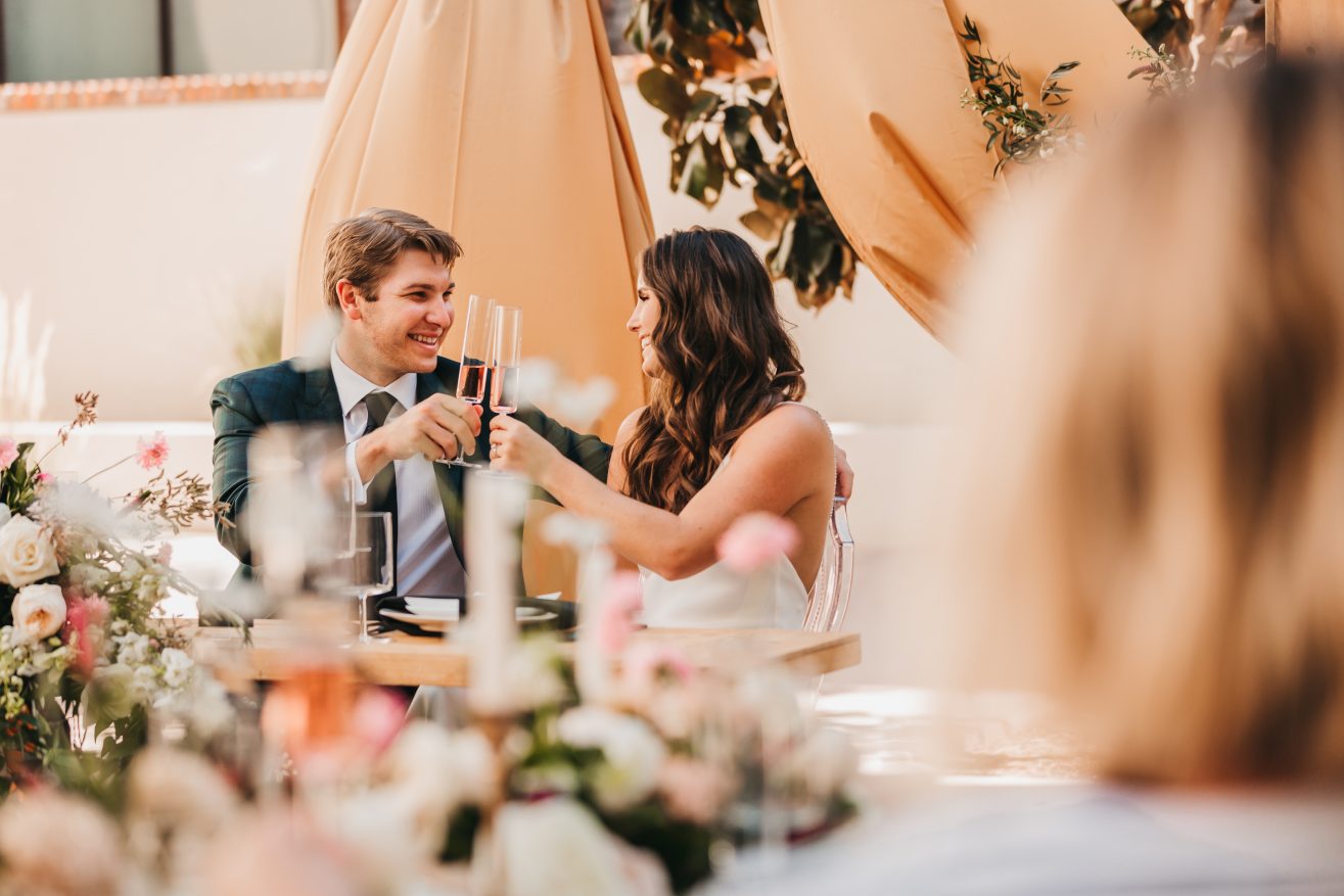 A bride and groom seated facing each other at a dinner banquet, holding their champagne glasses up together.