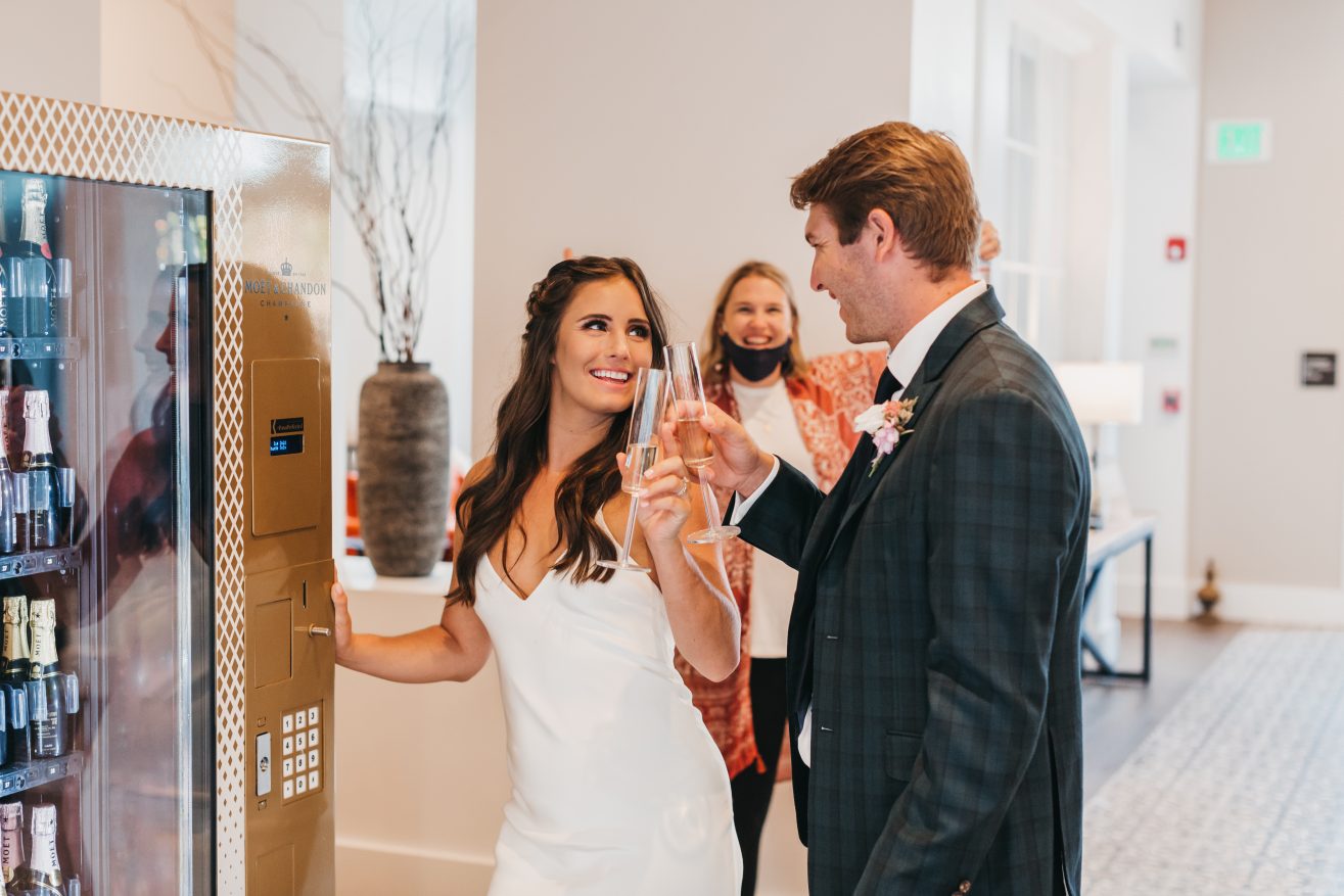 A bride and groom smiling at each other while holding champagne glasses and standing next to a vending machine. A happy woman is seen behind them with her arms up.