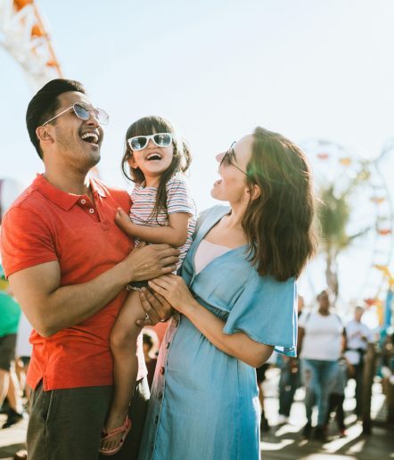 A man and woman holding a child all wearing sunglasses at a fair with a Ferris wheel in the background