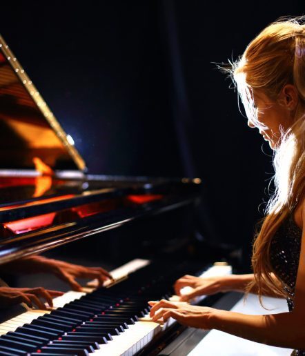 Side view of middle aged caucasian woman playing baby grand piano in a concert.She has long blond hair partially pulled back with a clip and shiny sleeveless dress.